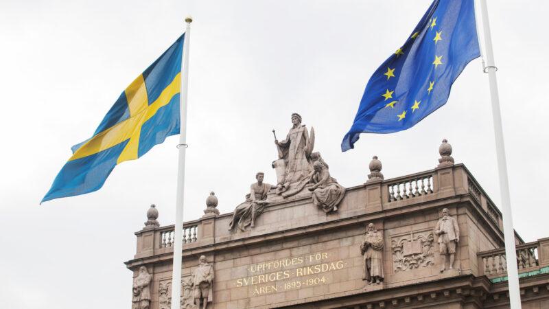Foto: Melker Dahlstrand /Sveriges riksdag Del av riksdagshuset, med en svensk flagga och en EU-flagga framför.